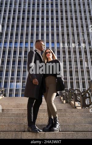 From below stylish multiracial couple in stylish outerwear hugging and standing on steps outside modern skyscraper on sunny day in city Stock Photo