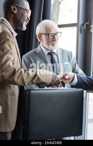 senior interracial businessmen giving briefcase to business partner in office Stock Photo