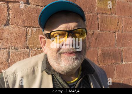 Outdoor portrait of smiling Caucasian senior man wearing blue cap and yellow sunglasses while standing against fulvous brick wall Stock Photo
