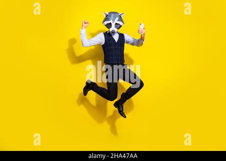 Full length photo of weird guy racoon mask jump raise fist up use device wear stylish trend vest isolated over yellow color background Stock Photo