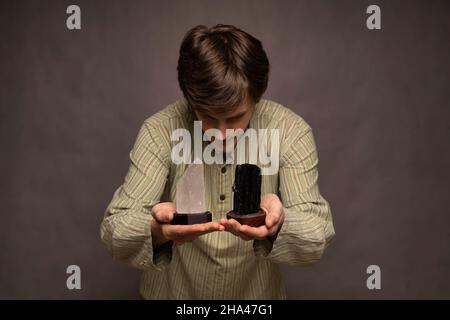 Young handsome tall slim white man with brown hair closely inspecting black and white crystals in striped shirt on grey background Stock Photo