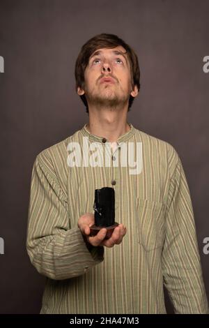 Young handsome tall slim white man with brown hair holding black crystal looking up in striped shirt on grey background Stock Photo