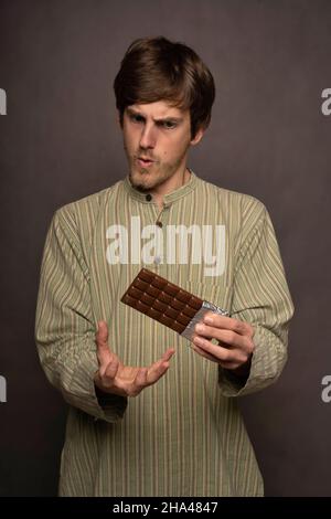 Young handsome tall slim white man with brown hair blown away by a chocolate bar in striped shirt on grey background Stock Photo