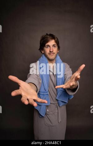 Young handsome tall slim white man with brown hair towel on shoulders reaching out with both arms in grey bathrobe on grey background Stock Photo