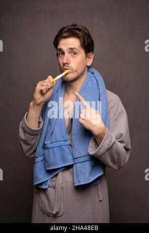 Young handsome tall slim white man with brown hair brushing his teeth pointing at the toothbrush in grey bathrobe on grey background Stock Photo