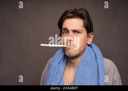 Young handsome tall slim white man with brown hair toothbrush in mouth looking confused in grey bathrobe on grey background Stock Photo