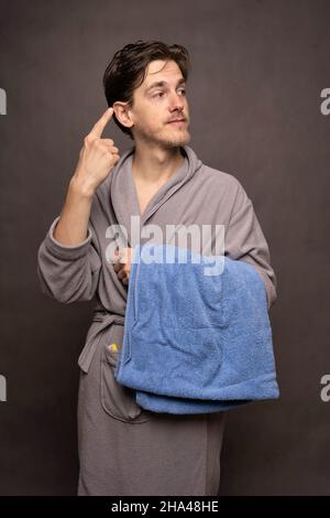 Young handsome tall slim white man with brown hair holding a towel pointing at his ear in grey bathrobe on grey background Stock Photo
