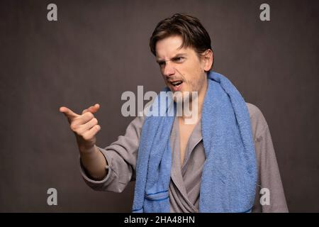 Young handsome tall slim white man with brown hair angrily pointing his finger with towel on shoulders in grey bathrobe on grey background Stock Photo
