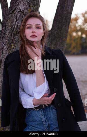 Long straight haired white european woman in dark coat near the tree Stock Photo