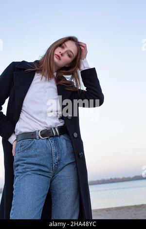 Long straight haired white european woman touching her head in dark coat in autumn on the beach Stock Photo