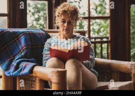 pretty middle age woman relax at home sitting on an armchair reading a book enjoying indoor leisure activity. adult female people comfortably sit down on the couch read novel and enjoy comfort Stock Photo