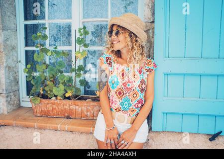 beautiful woman in sunglasses and knit hat standing against closed glass window of home. young caucasian woman posing against residential house. hipster tattooed woman posing in sunglasses and hat Stock Photo