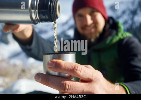 Portrait of man with beard out of focus pouring hot tea from thermos in the small cup close up against winter mountain background. Stock Photo