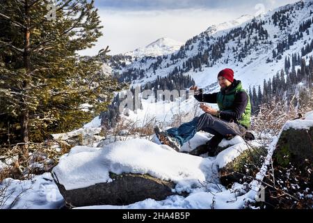 Man with beard in red hat pouring hot tea from thermos in the spruce forest against winter mountain background. Stock Photo