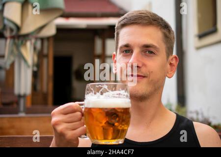 Young handsome white man with brown hair in black undershirt is happy with pint of beer in his hand on pub terrace Stock Photo