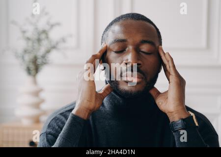 Stressed African home office worker sits at desk touching temples while eyes closed Stock Photo