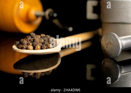 Lot of whole spicy black pepper in a wooden spoon with pepper mill with aluminum mortar isolated on black glass Stock Photo