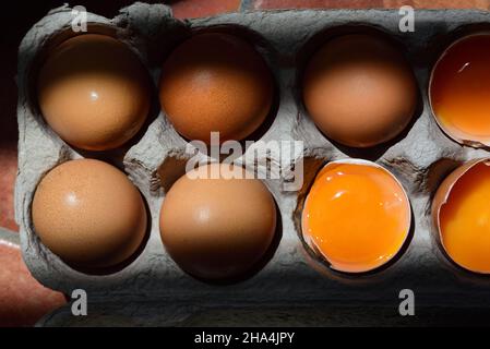 In a cardboard egg box are several eggs, some of which are still intact and others open with the yellow egg yolk, photographed from above, in a play o Stock Photo
