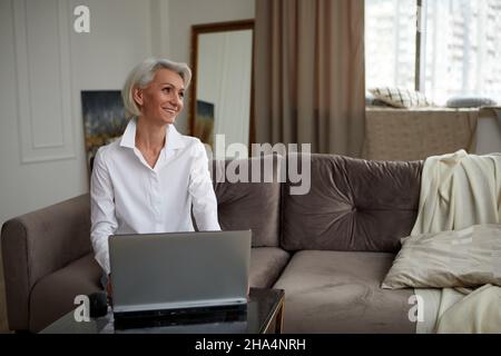 Happy 50s older mature middle aged adult woman holding laptop using computer sitting on couch at home. Smiling elegant senior grey-haired lady spendin Stock Photo