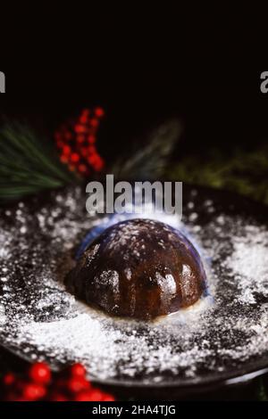 close up of a flaming Christmas pudding on a plate with icing sugar and surrounded by seasonal foliage. Stock Photo