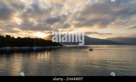 greece,greek islands,ionian islands,kefalonia,fiskardo,morning mood,partly cloudy sky,view of the sunrise over the harbor basin,gray cloud cover,ithaca in the background,small fishing boat is just entering the harbor,wide-angle view,dynamic sky over gray golden and silver shimmering sea Stock Photo