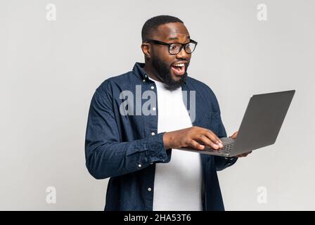 Excited young African American man standing isolated on grey wall holding laptop computer and typing message, chatting with friend on social media online, using dating app, enjoying new application Stock Photo