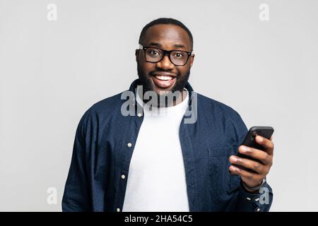 Cheerful African-American young man in casual shirt and glasses holding smartphone, testing mobile app, surfing in networks, scrolling news feed in social networks, isolated on grey background Stock Photo