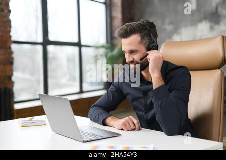 Concentrated young guy in smart casual wear is using headset and laptop for online communication, supporting, selling. Guy sitting at the office desk Stock Photo