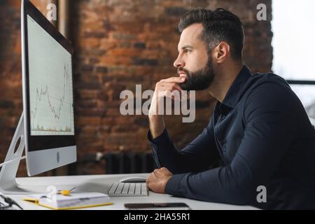 Focused and concerned man in smart casual shirt looking at laptop screen. Serious caucasian guy sitting at the desk and feeling doubts, while solving difficult tasks Stock Photo