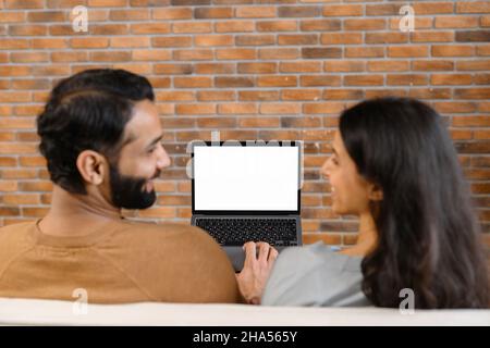 Back view of the young Pakistani couple using laptop while sitting on the sofa at home. Spouses watching something on laptop with empty screen, mock-up, copy space, template Stock Photo