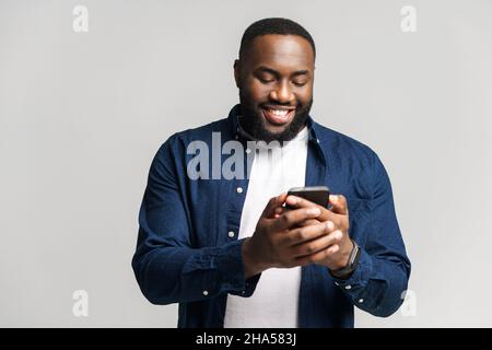 Optimistic African-American man wearing casual shirt texting on the smartphone, texting, smiling black guy enjoying using new mobile app isolated on grey Stock Photo