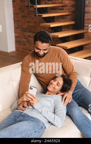 Indian couple with smartphone on the couch spending leisure time in social networks, scrolling feed, watching videos. Woman laying at the knees of her boyfriend Stock Photo