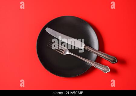 Minimalism style. Empty black plate and a cutlery set on red paper background. Stock Photo