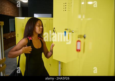 Beautiful young African athlete woman in black stylish sportswear opening locker in gym wardrobe, putting her bag inside it before fitness training Stock Photo