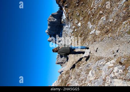 middle-aged woman,handbag,anorak,taken from behind on the passamani circular route,in the background the summit cross of the western karwendelspitze (2385m),mittenwald,germany,bavaria,upper bavaria,blue sky,karwendel mountains, Stock Photo