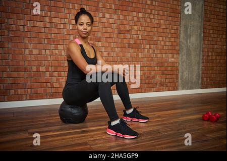 Beautiful African athlete woman in stylish black activewear sitting on a medicine ball and looking at camera at gym studio with red bricks wall Stock Photo