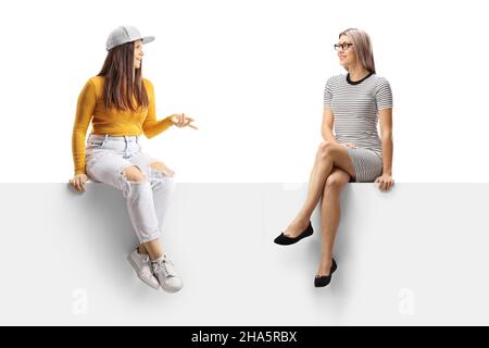 Two young women sitting on a blank panel and having a conversation isolated on white background Stock Photo