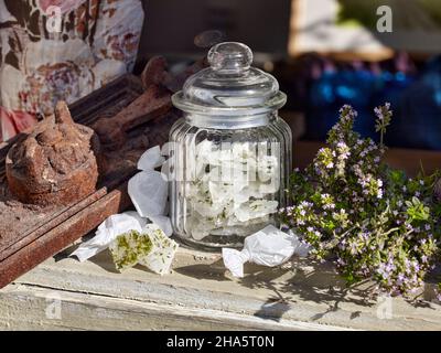 candy jar with lid,filled with thyme candies,on an old wooden window sill next to a bouquet of thyme and candies wrapped in paper Stock Photo