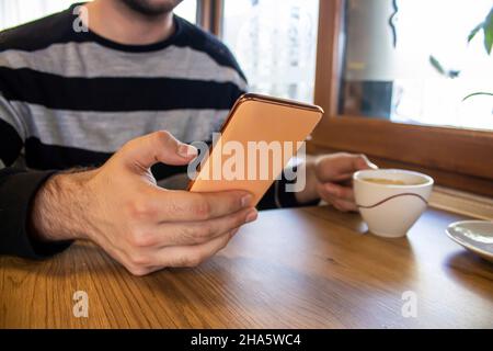 Man using smartphone while drinking coffee, young man sitting in a cafe and holding mobile phone. Stock Photo