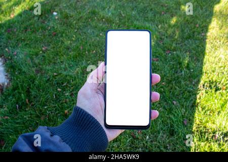 Hand holding smartphone, mockup image of man hand holding mobile phone with blank white desktop screen while walking on green grass. Stock Photo