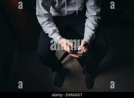 Close up of teen boy in white sweater texting on phone in dark. Stock Photo