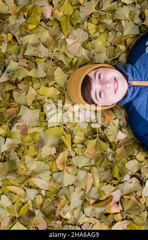 Overhead of happy boy laying on a pile of yellow leaves on fall day. Stock Photo