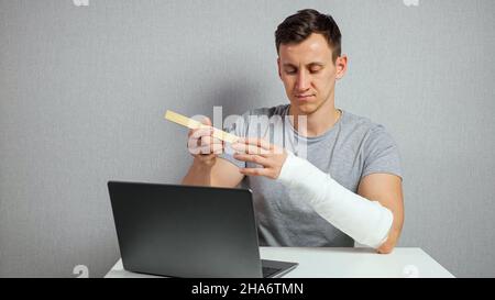 Young man scratches arm in gypsum band working on laptop Stock Photo