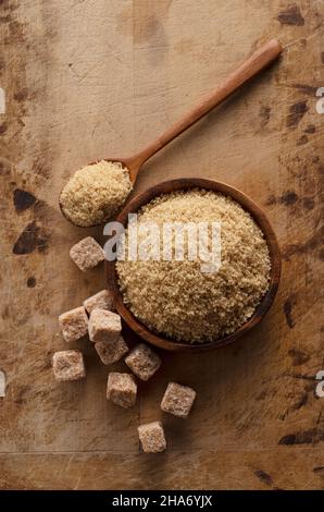 A wooden plate set against an old wooden background, brown sugar in a spoon, and a sugar cube next to it. View from directly above. Stock Photo