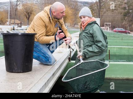 Man owner of sturgeon farm with female worker inspecting fish in tank outdoors Stock Photo