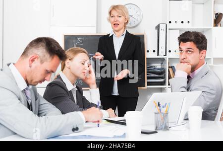 Mature businesswoman standing near blackboard in office, expressing dissatisfaction with work of colleagues Stock Photo
