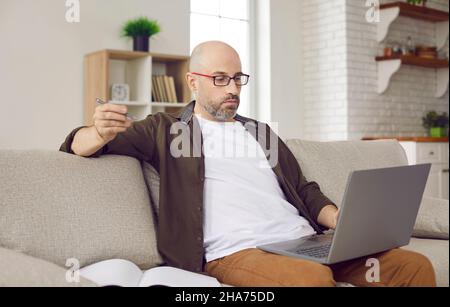 Portrait of adult male freelancer working remotely at home using laptop and notebook. Stock Photo