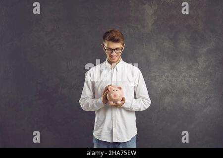 Smiling man standing on gray background looks at piggy bank in his hands with smile on his face. Stock Photo