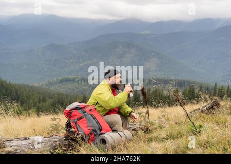 a bearded Caucasian male tourist on vacation while hiking in the mountains sits on the trunk of a fallen tree and drinks tea or coffee from a thermos Stock Photo