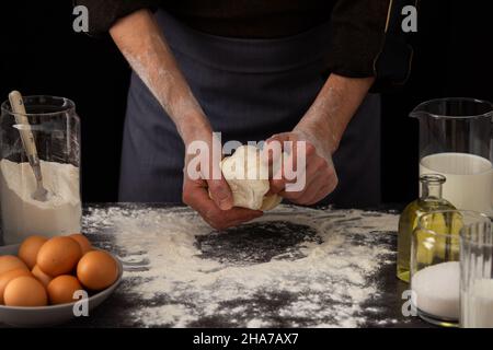 A man baker kneads dough on the table, milk in a jug and flour, chicken eggs in a plate and sugar and butter, male hobby for cooking Stock Photo